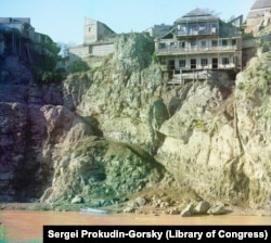 A clifftop house in Tbilisi with what appears to be a precarious pathway leading down to the river and a boat. A pipe on the right is dribbling wastewater into the Mtkvari River.