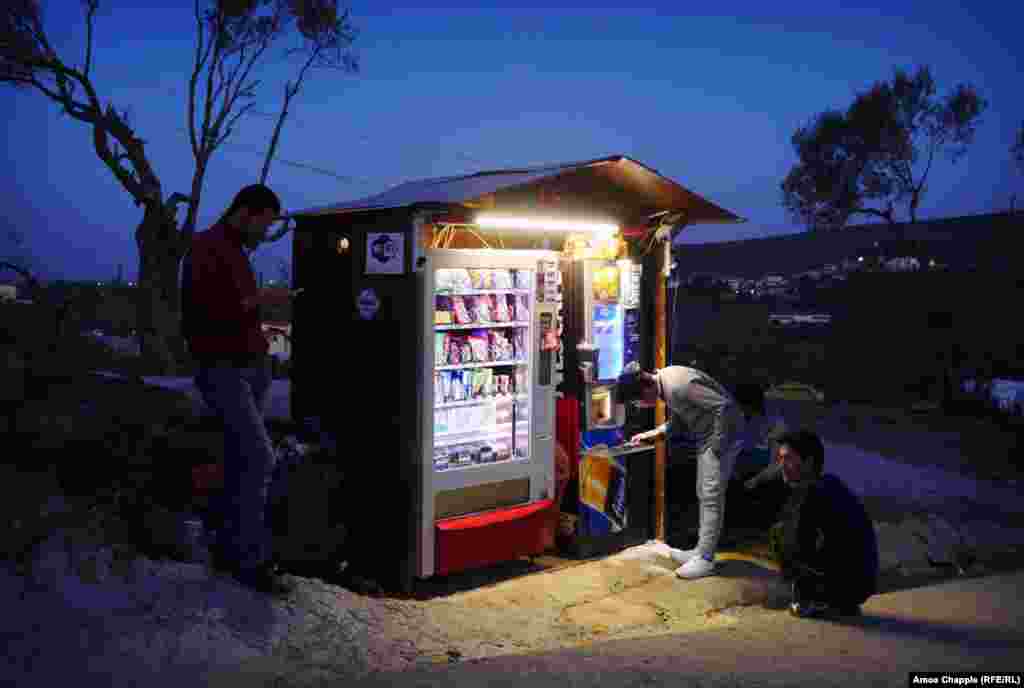 Dusk&nbsp;on the edge of the camp, where migrants charge their&nbsp;cellphones&nbsp;and use the free Wi-Fi. In the background, the lights of&nbsp;Moria&nbsp;village&nbsp;twinkling to life&nbsp;mean&nbsp;locals will be gathering in the&nbsp;tavernas&nbsp;for seafood and a drink&nbsp;and, as always, some heavy discussion.