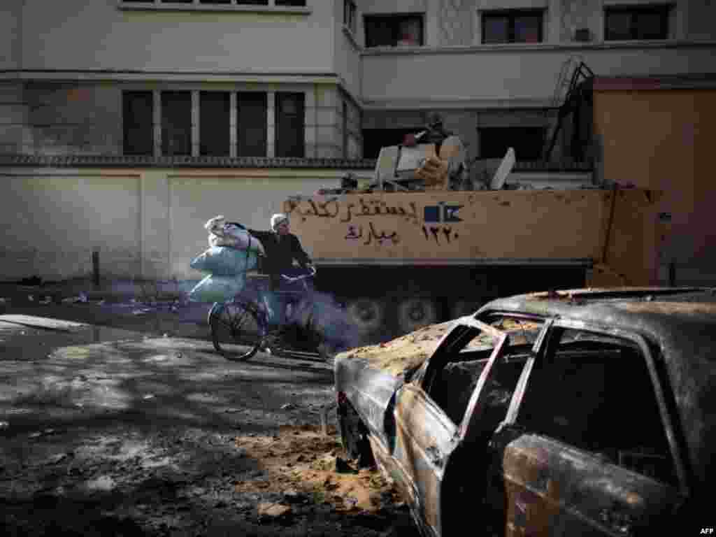 A man passes between charred remains of a looted car and an Egyptian Army armored personnel carrier.